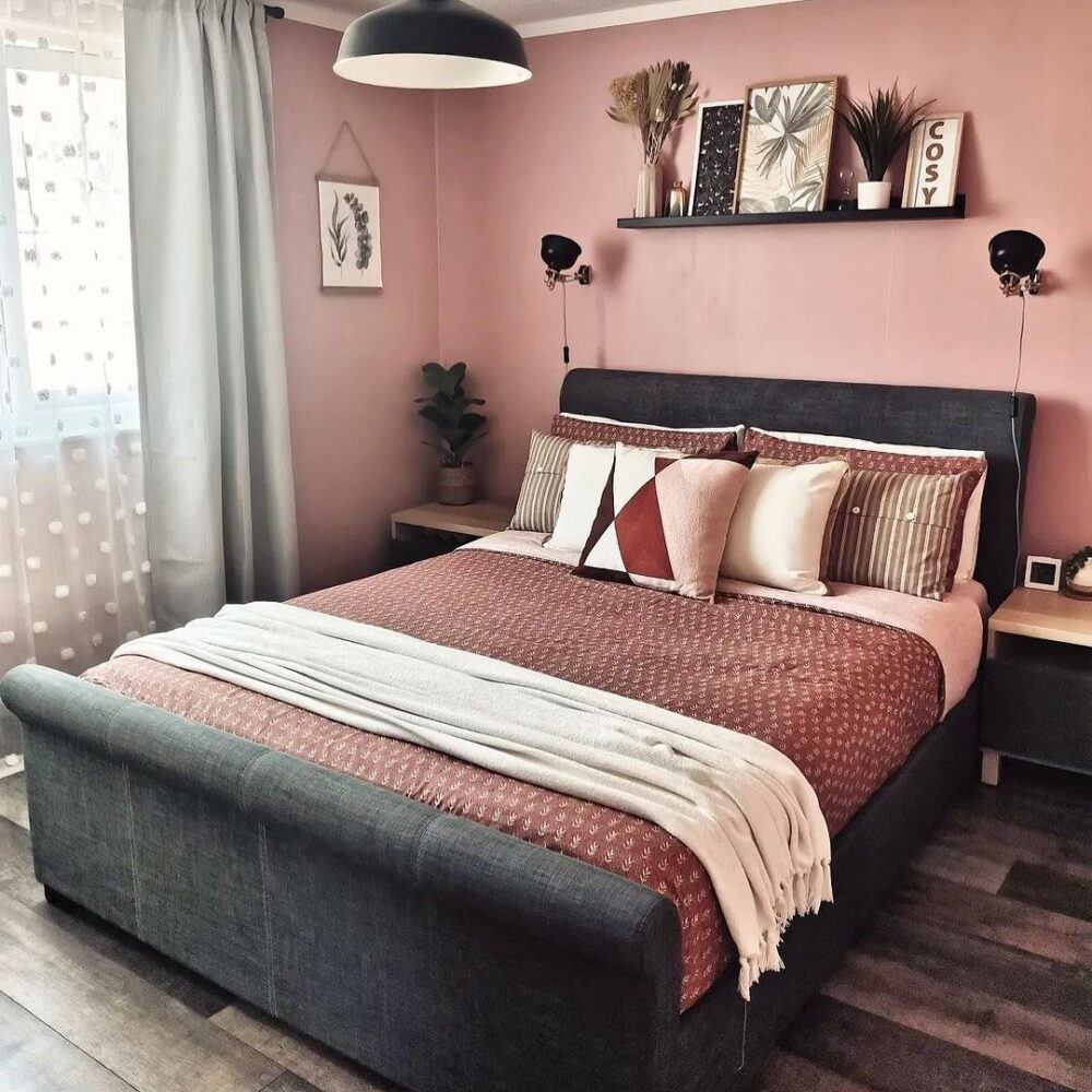 Image shows a burgundy and pink bedroom with a black bed frame. There are light pink sheets with burgundy throws and pillows. White scatter cushions adorn the bed and there's a deep, dark stained wood floor.