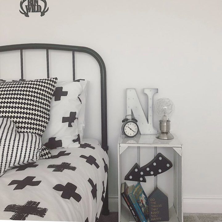 A white and black space, with an open concept bedside table. Inside are nestled a collection of books, while atop it rests an alarm clock and a lamp. There is a black metal bed, with varying textures printed on the pillowcases and blankets.