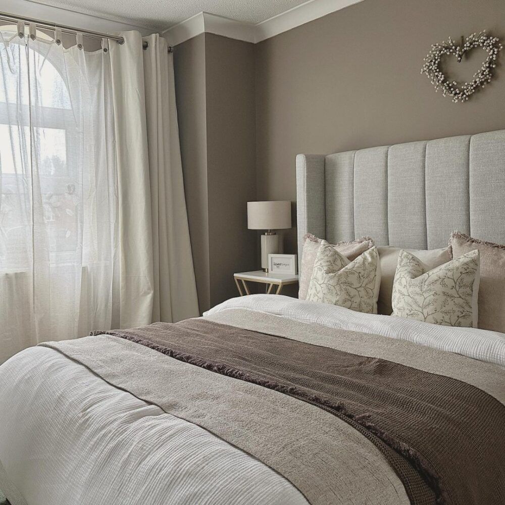 A grey upholstered bed, styled in a soft brown and grey bedroom with sandy tones and soft furnishings.