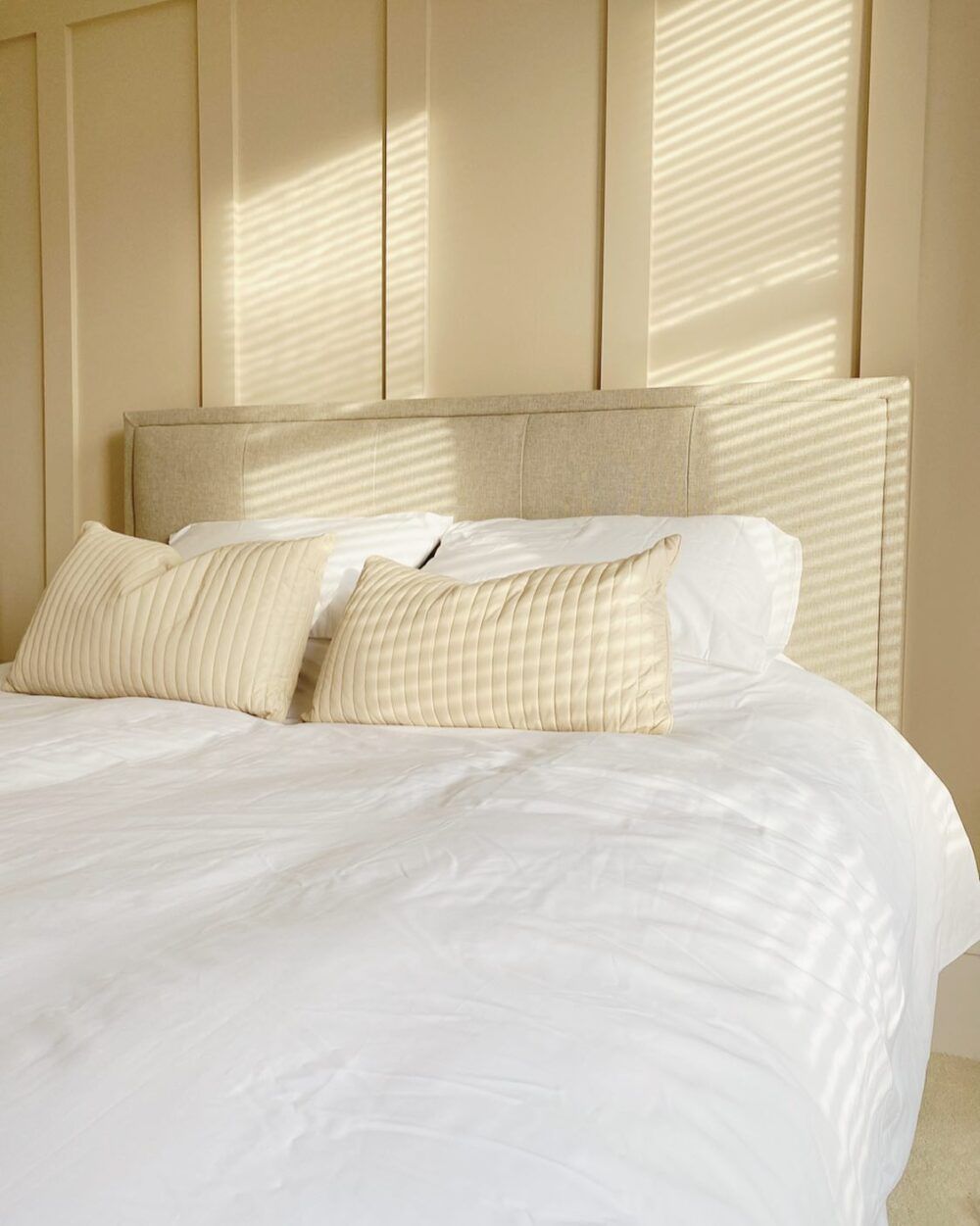 A bed with white bedding, two yellow pillows, and a light beige headboard. Set against a soft yellow wall with panelling.