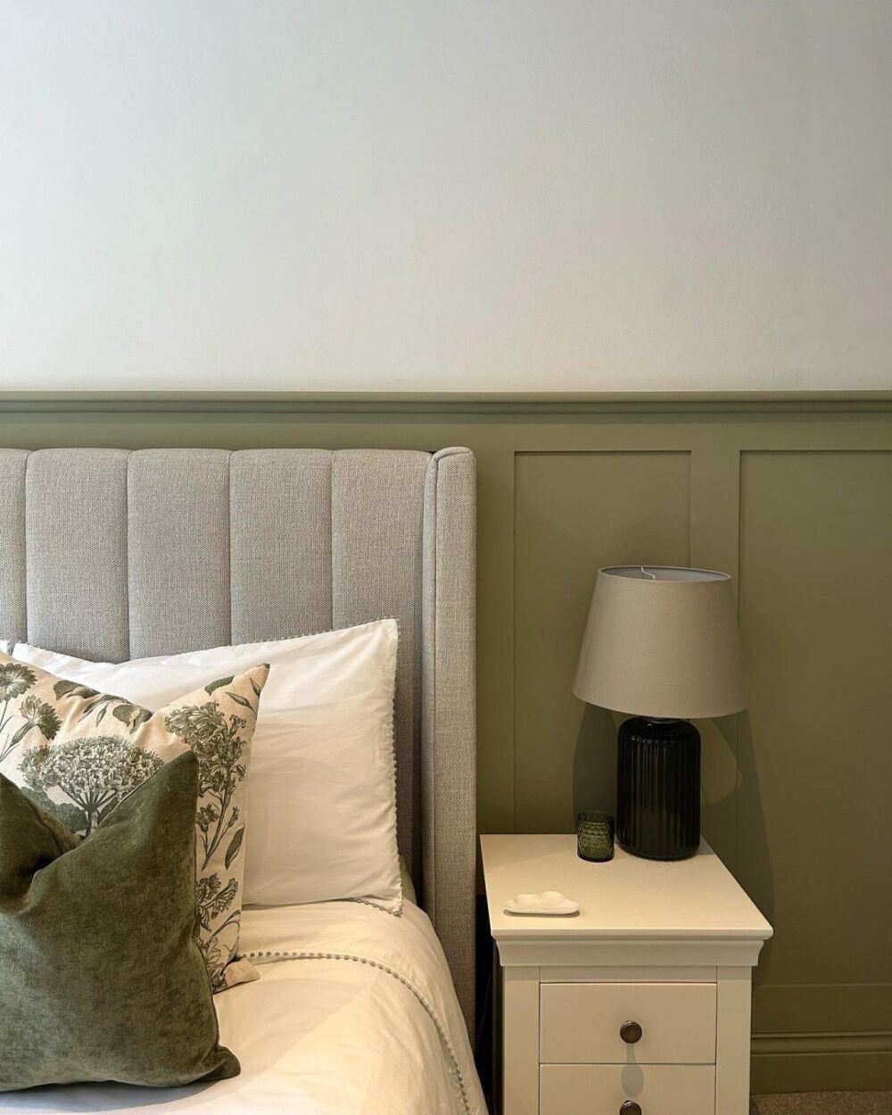 A grey upholstered bed frame, set against a olive green panelled bedroom wall.