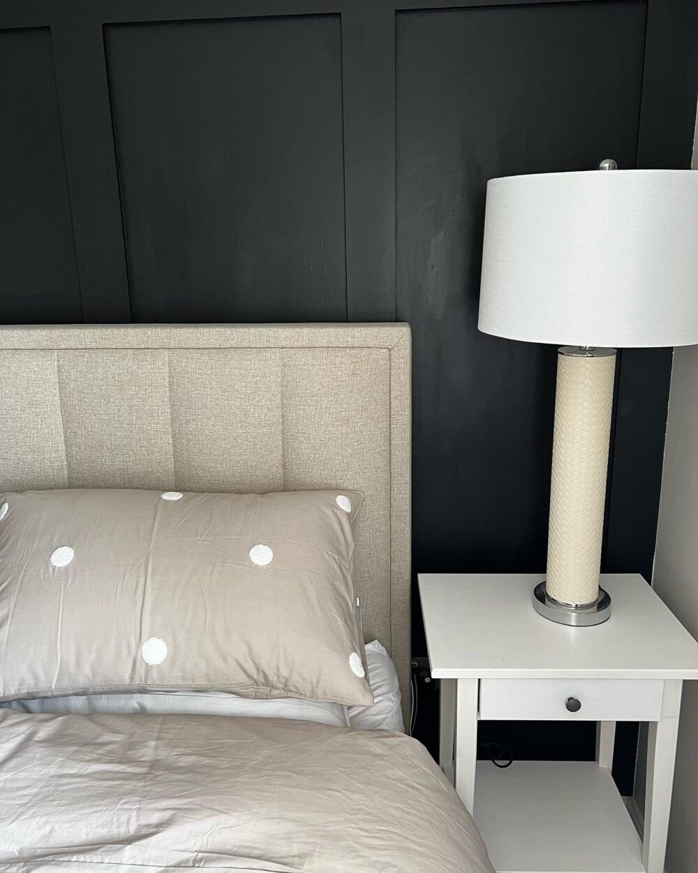 The corner of a beige headboard behind beige bedding. The bed is next to a small white bedside table with a white lamp, and against a black wall.