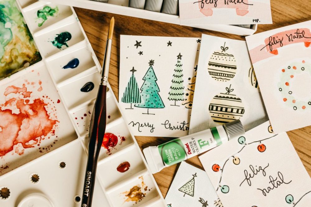 A collection of hand-painted Christmas cards scattered on a wood table with a paint palette on the left side of the image.