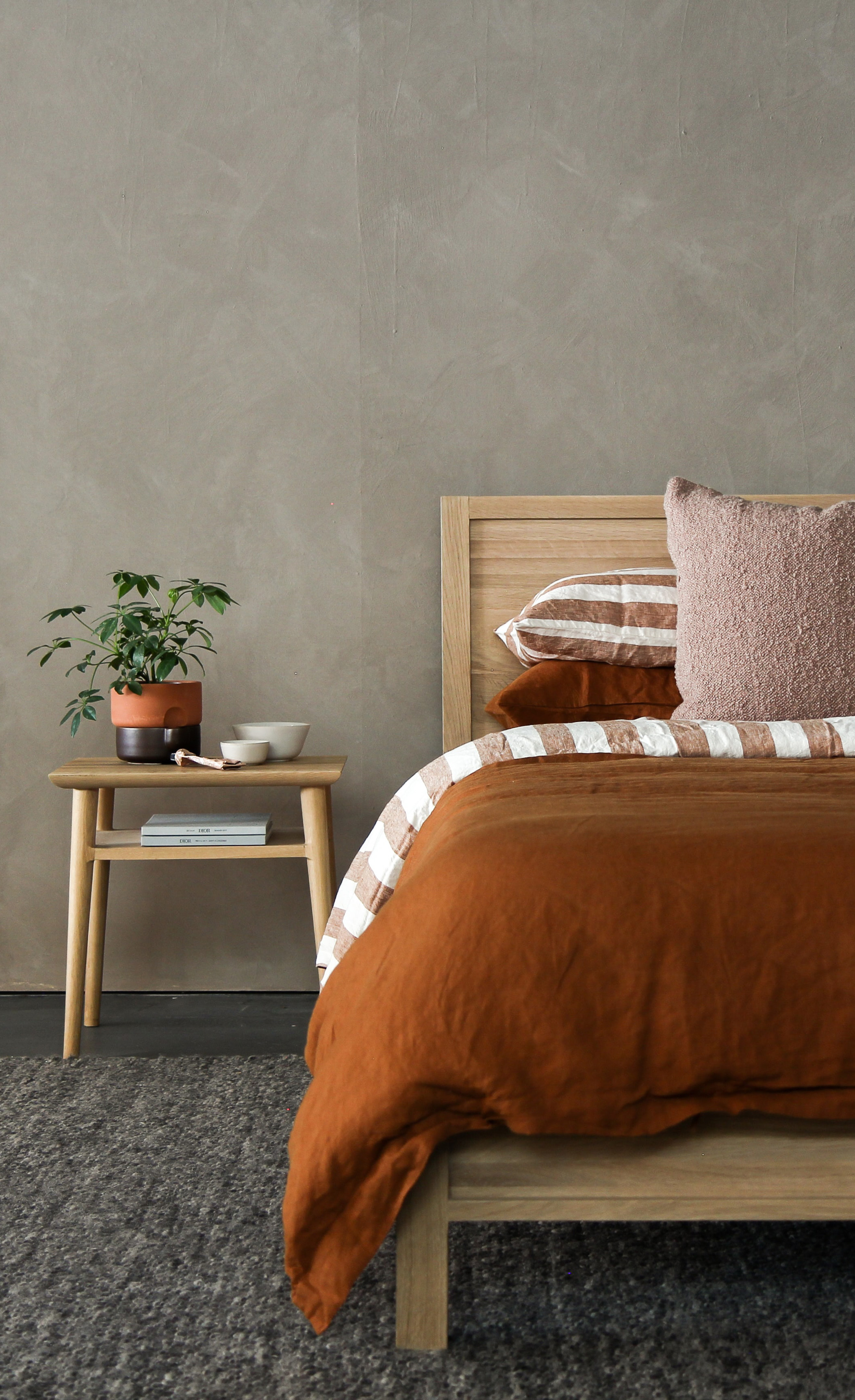 wooden bed with mattress and bedding on top. In the corner, there's a small retro 70s style table with a plant on top.