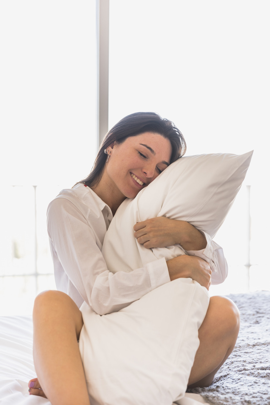 A young woman with straight hair sitting on bed hugging her white pillow, surrounded by white