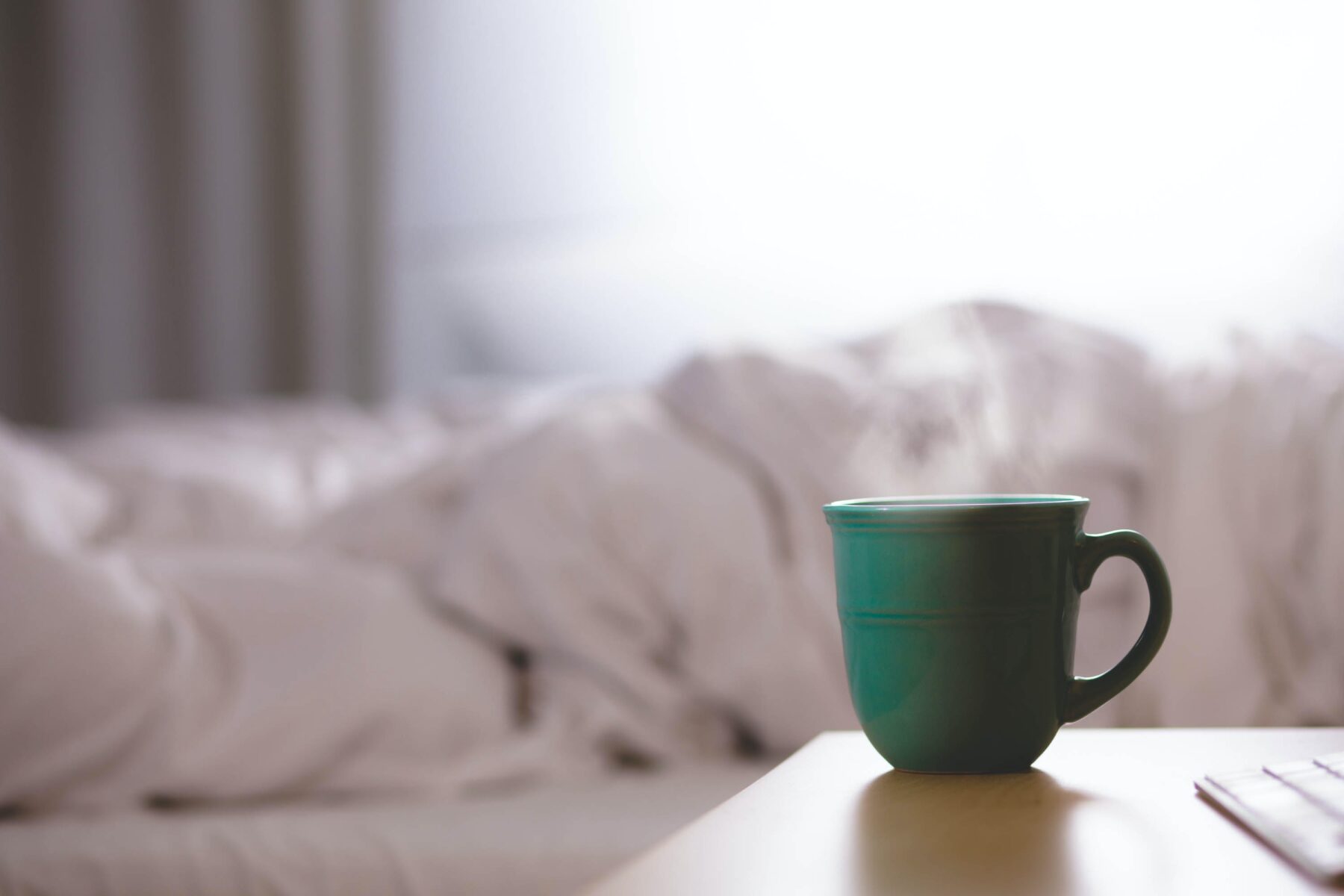 A green cup of tea steaming in a bedroom, on a wooden bedside table. In the background, messy white sheets and grey curtains can be seen.