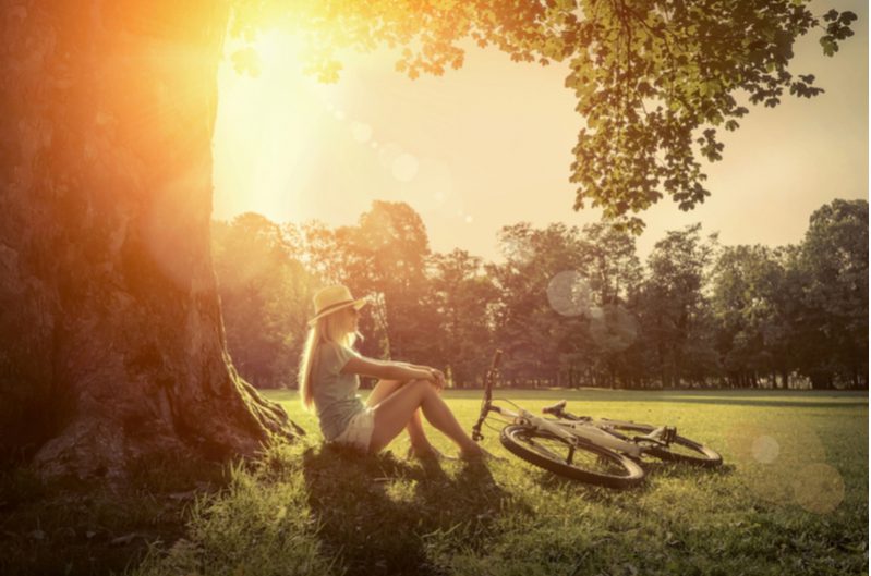 Someone sits at the foot of a tree with a sunhat on and a bike nearby, relaxing in the sun.