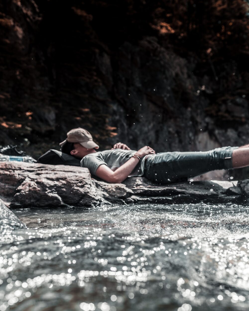 A person sleeping mid-hike with legs elevated, a cap over their face, and water running nearby from a river.