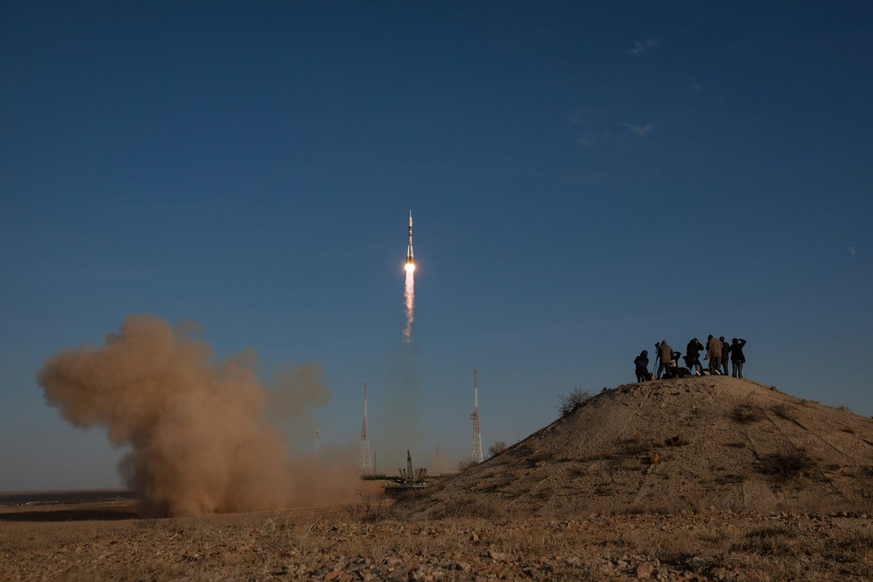 Image of a nasa rocket taking off at dusk with a group of people watching from a hill in a sandy, desert like environment