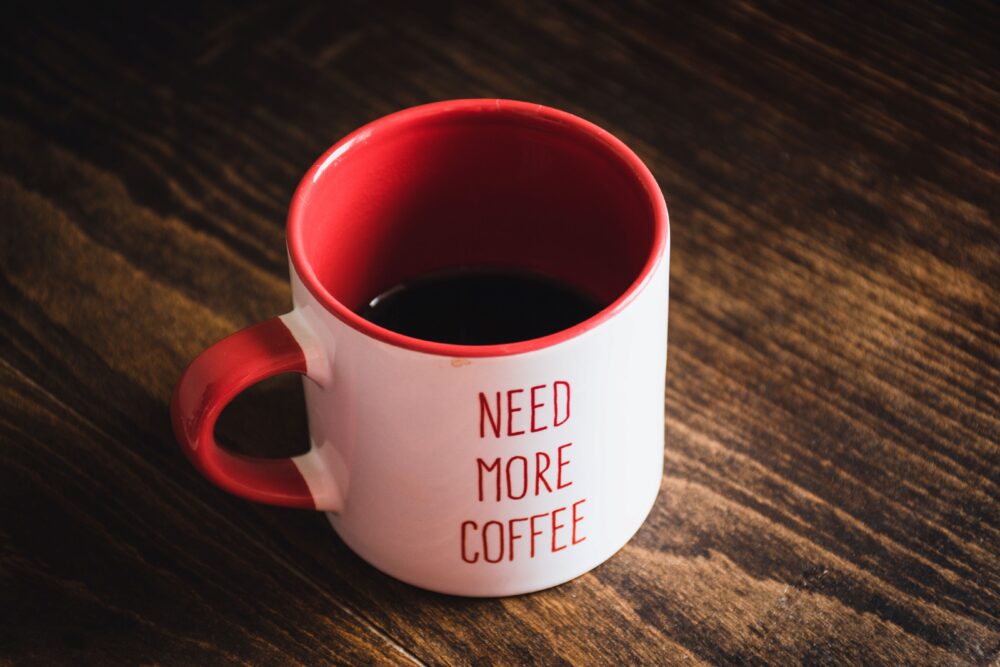 A white and red mug on a wooden table, half-filled with black coffee. The words "NEED MORE COFFEE" are printed on the mug.