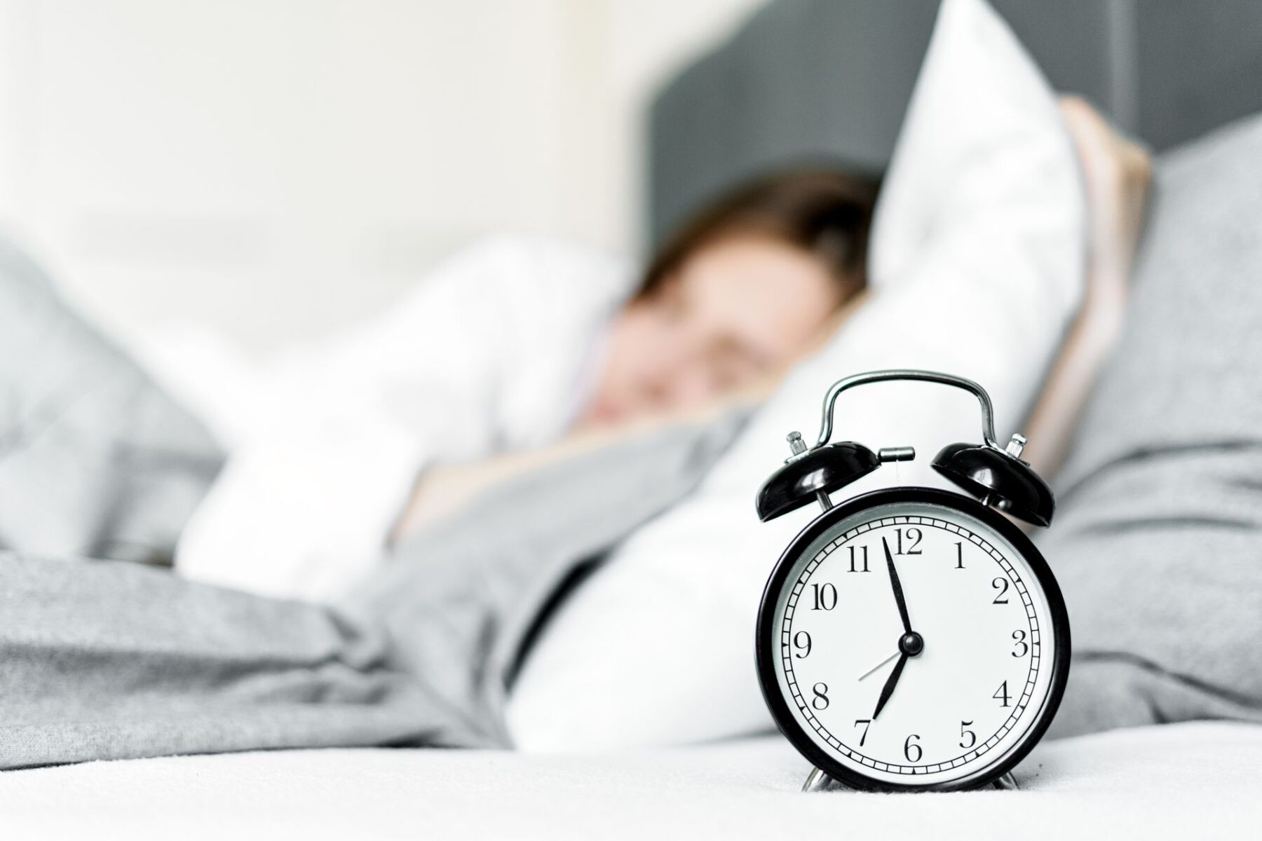 A retro alarm clock sits by a sleeping woman in a cosy bedroom setting.