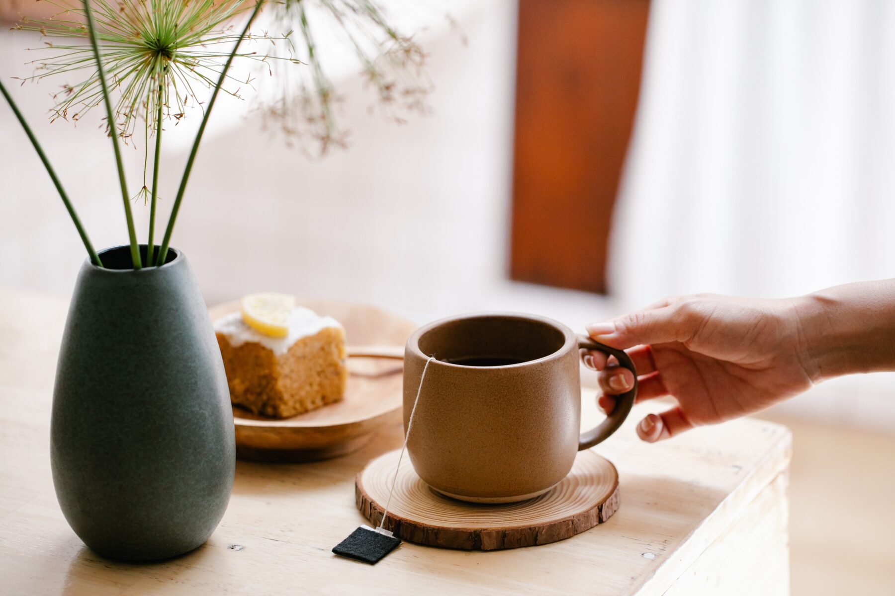 A cup of tea sits on a wooden coaster, with a slice of cake and a flower vase in the background.