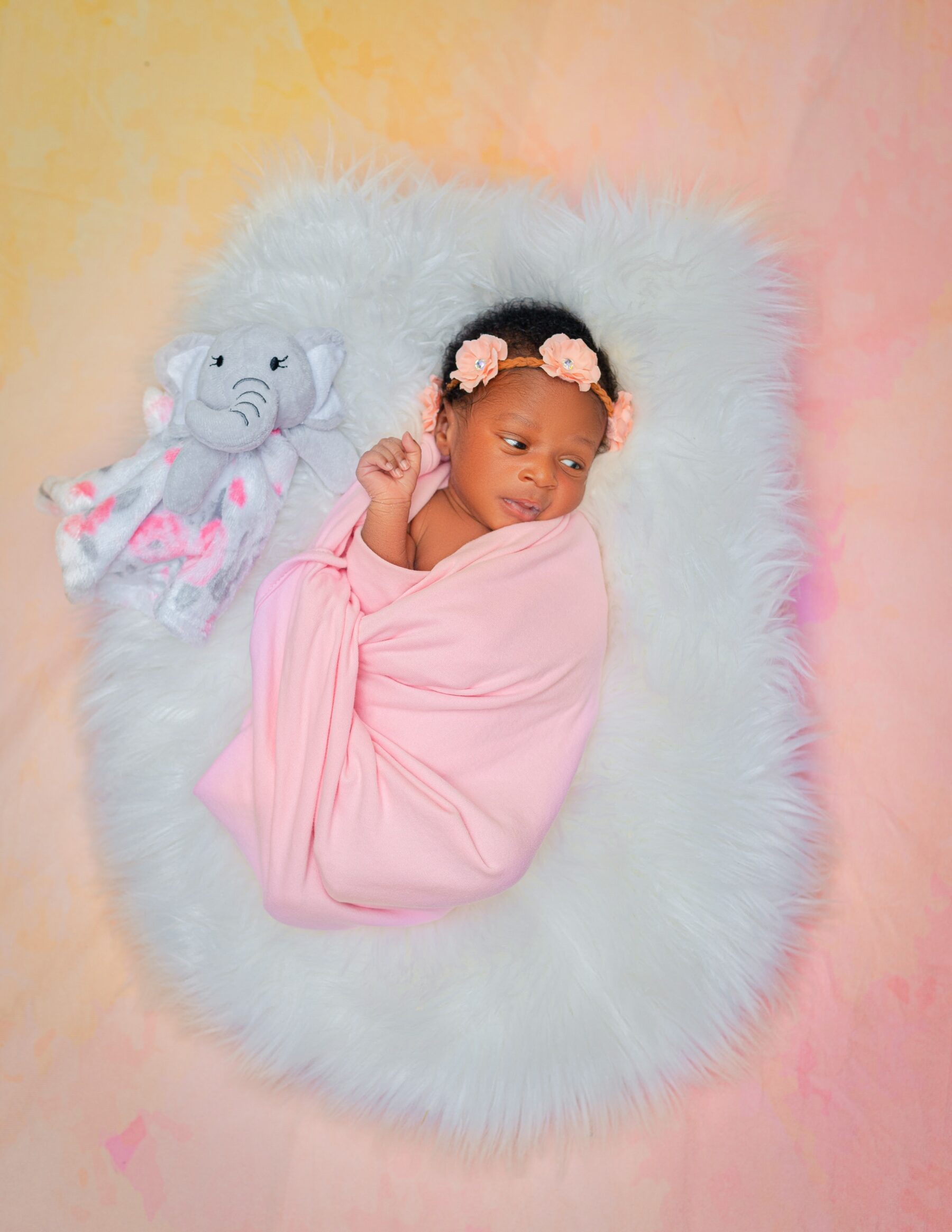 a baby swaddled in a pink blanket with a pink bow in her hair, next to a soft toy and on a soft fluffy grey bed.