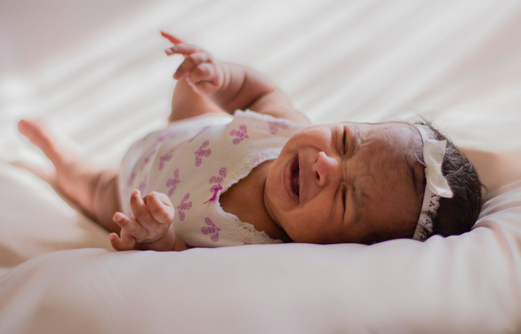 A baby cries on a white blanket, while wearing a white and pink flower print onesie and a headband with a bow.