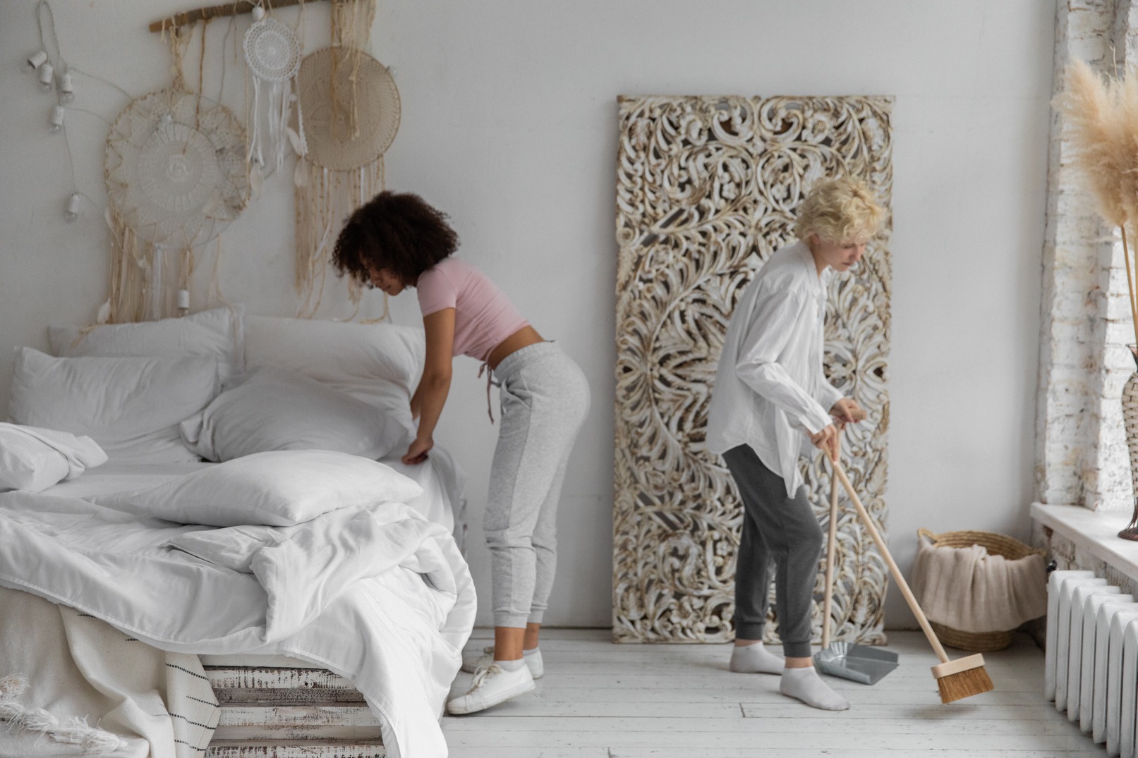 A couple are tidying a white bedroom together. One of them is sweeping the wood floors, while the other is changing the bedsheets.