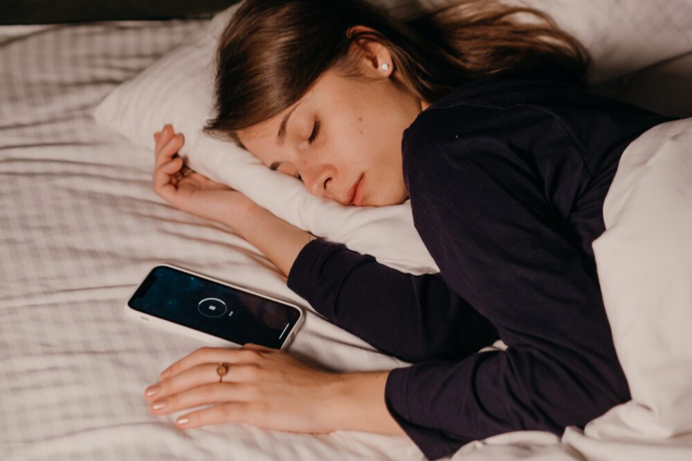 Woman in black shirt sleeping in white bed with phone next to her