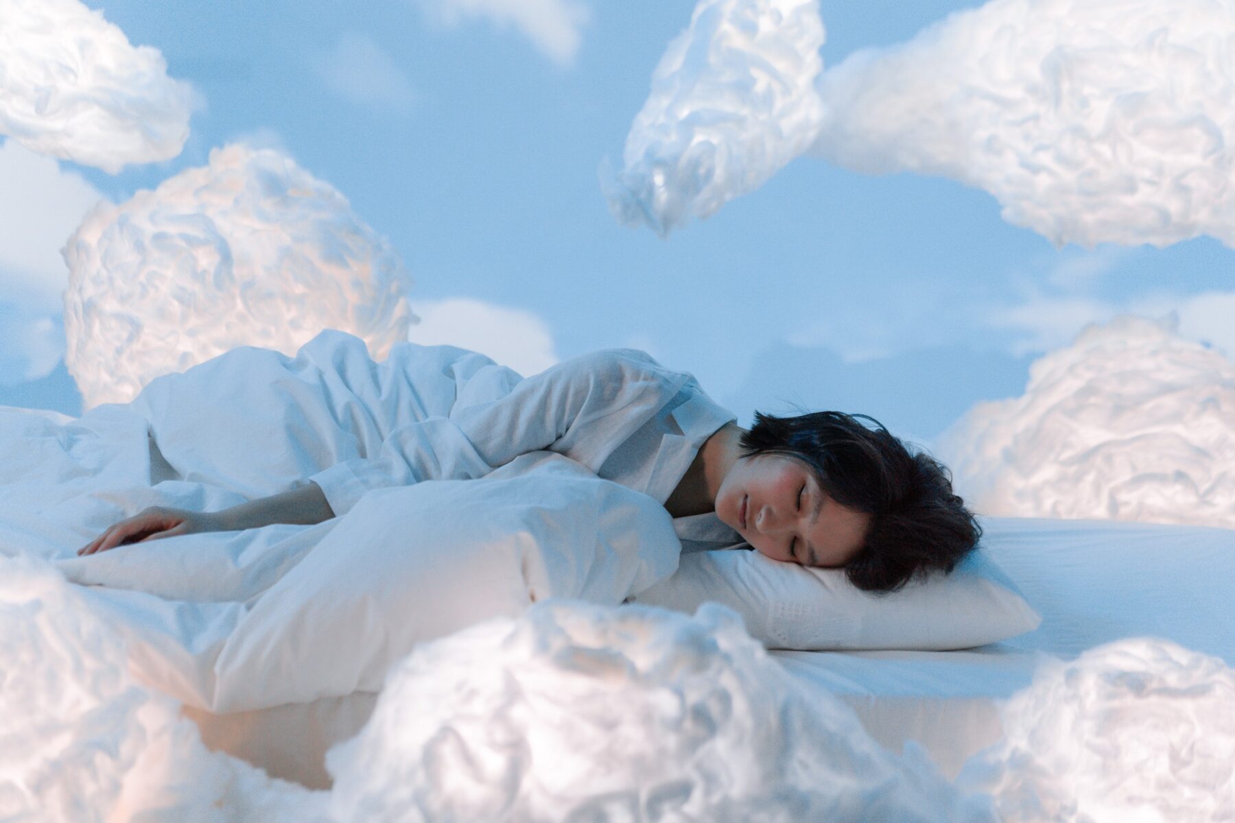 A person sleeps on a white sheet surrounded by fluffy clouds and a blue backdrop.