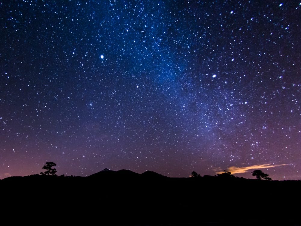 A view of the night sky, with purple and blue hues, and stars, backlighting a blackened landscape on a clear night.