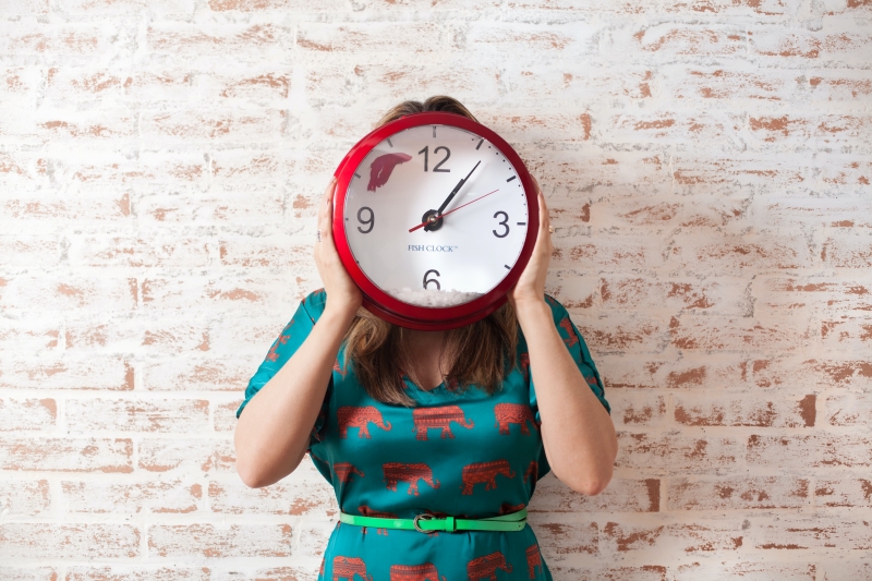 A woman in a green and red dress standing against a whitewashed brick wall holds a red clock to their face