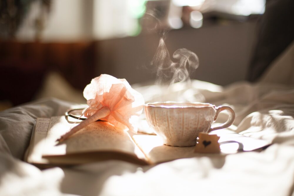 A cup of steaming tea in an ornate white cup, on a book next to a pink flower, set on a bed.